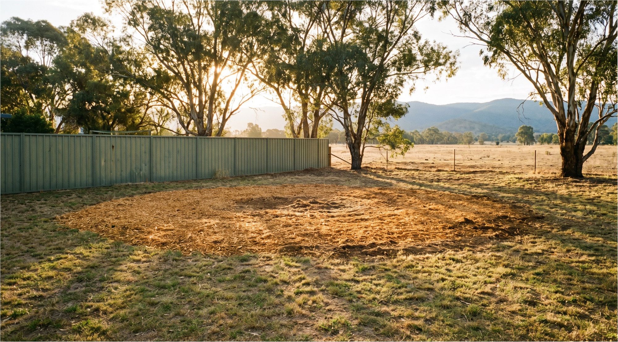 A wide circle of fresh wood chip mulch in a country backyard beside a Colorbond fence, with gum trees and hills behind